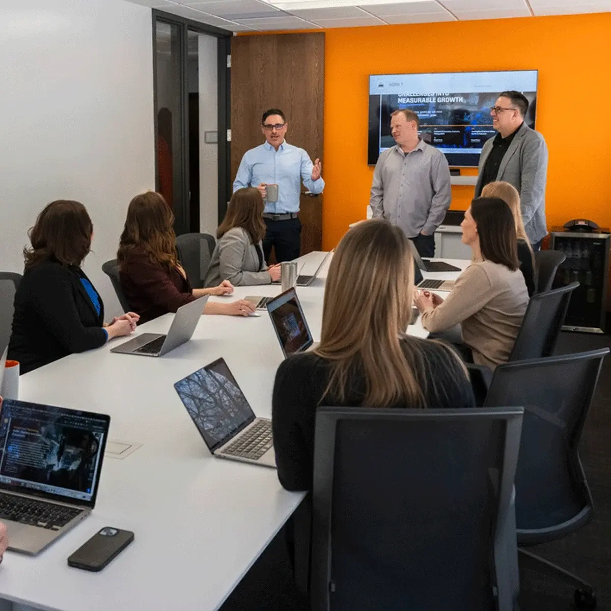 A presenter addresses colleagues seated around a conference table in an office meeting room with an orange wall.